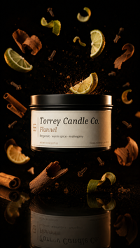 A jar candle from Torrey Candle Co. labeled 'Flannel' is centered against a black background with floating warm spices, cinnamon sticks, cloves, and citrus slices around it.